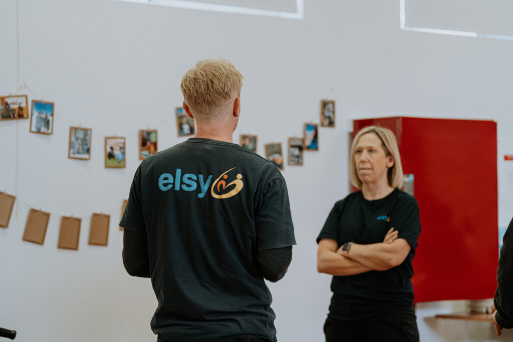 A blonde woman in a black t-shirt with her arms crossed stands to the right of the man.