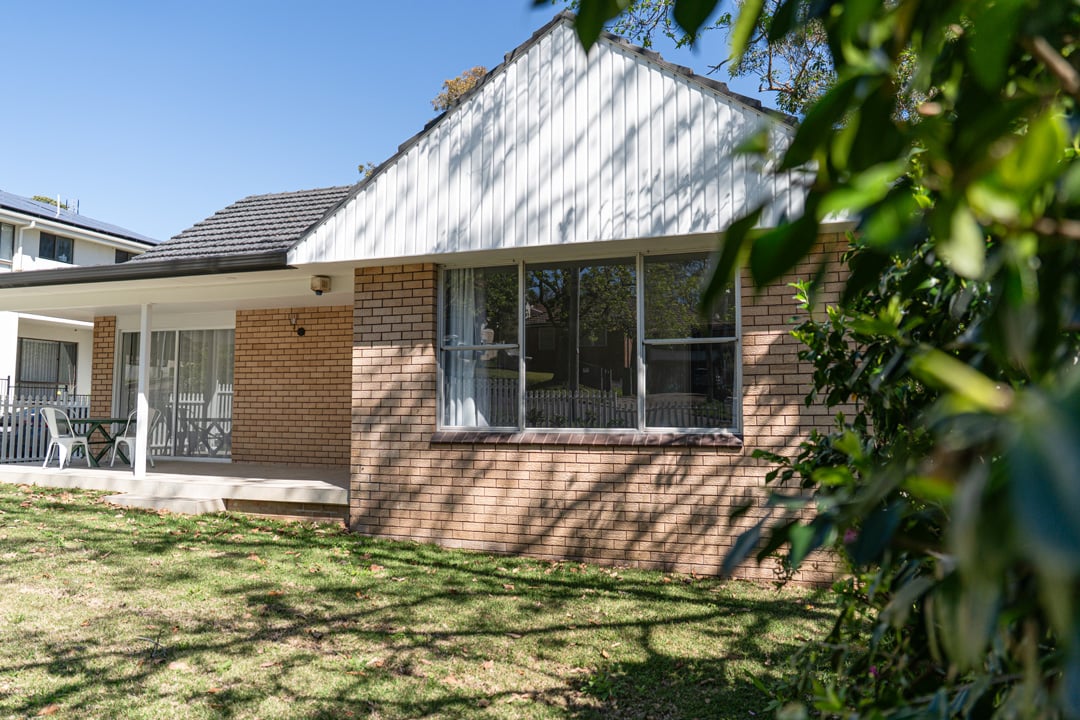 Exterior view of a single-storey Australian suburban house with light brown brick walls and a white vertical-cladded gable.