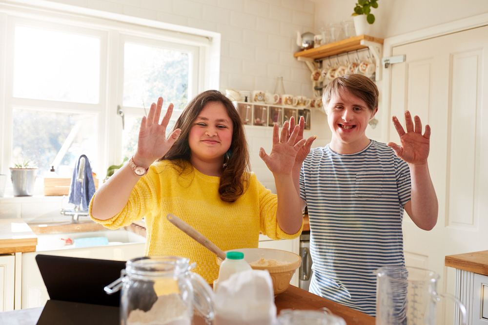 Two teenagers, a girl and a boy, standing behind a kitchen counter in a brightly lit kitchen.