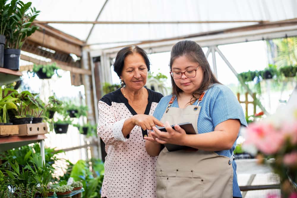 A greenhouse or nursery, showing two women looking at a digital tablet.